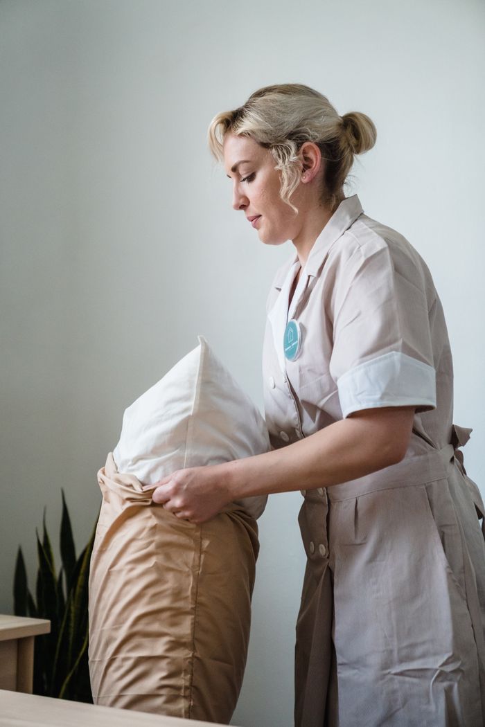 who-we-are A hotel housekeeper in uniform is straightening pillows in a neatly arranged guest room.